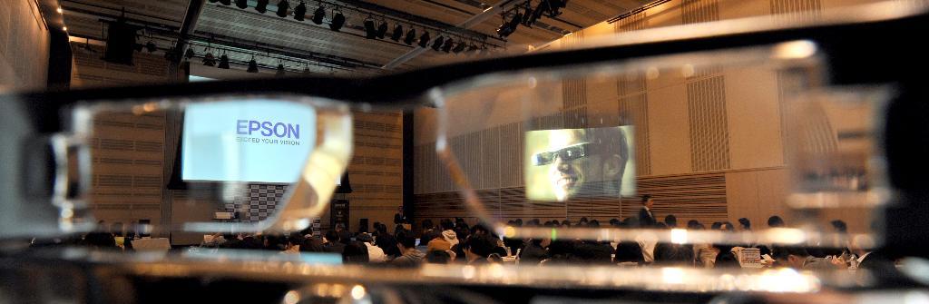 エプソンが発表したメガネ型情報端末「ＭＯＶＥＲＩＯ（モベリオ）ＢＴ－２００」。右側レンズに映し出された映像をモベリオをかけた際に大画面で体感できる＝２８日、東京・大手町（寺河内美奈撮影）