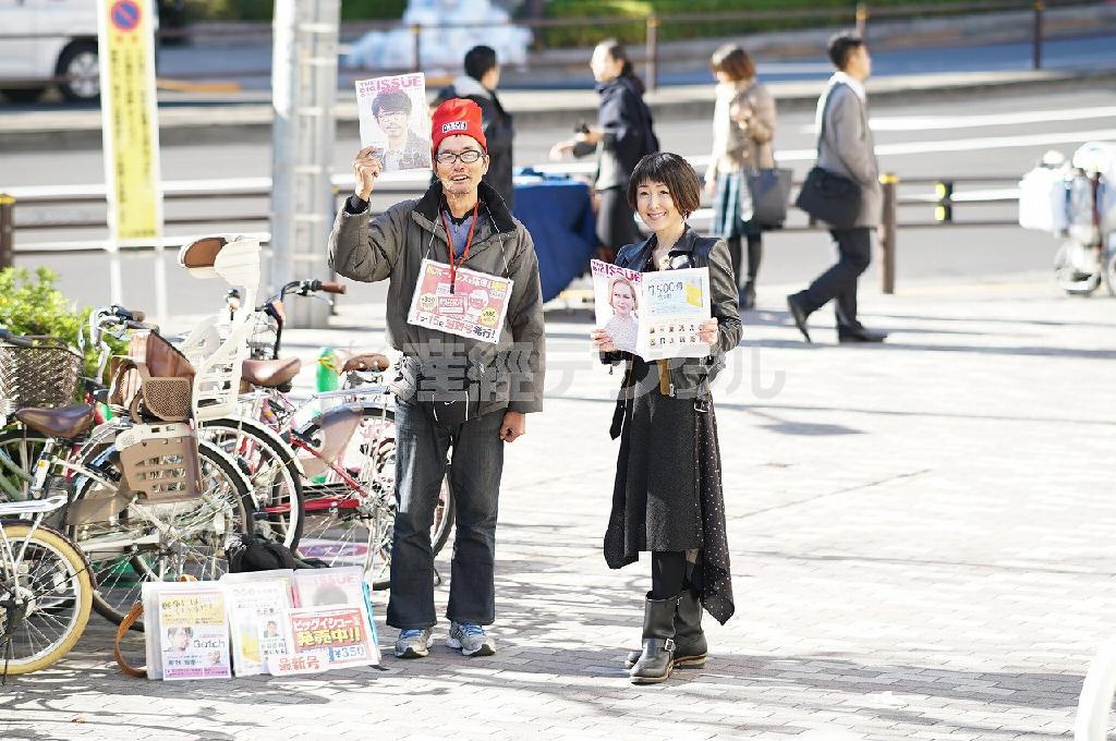 人通りの多い交差点に立つ雑誌「ビッグイシュー日本」（１冊３５０円）の販売者、菅野さん（左）と一般社団法人「Ｇｅｔ＿ｉｎ＿ｔｏｕｃｈ」理事長の東ちづる＝東京都文京区・本郷三丁目の駅前交差点（山下元気さん撮影）