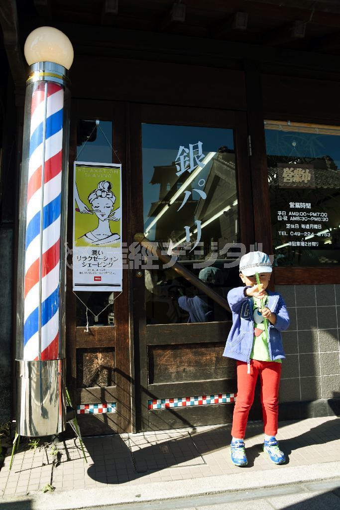 店名「銀パリ」の書体や、ドアに貼り付けられた赤青白のタイルが昔ながらの理髪店から、散髪を終えた男の子が出てきました。少し照れくさそうに持っていたお菓子で顔を隠しながらもポーズ。手前の理髪店の動く看板と木の扉や壁の露出が違い過ぎ、手前に合わせると木目が出なかったので、少し明るめに露出決定しました＝２０１４年１１月４日、埼玉県川越市（須藤夕子さん撮影）