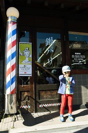 店名「銀パリ」の書体や、ドアに貼り付けられた赤青白のタイルが昔ながらの理髪店から、散髪を終えた男の子が出てきました。少し照れくさそうに持っていたお菓子で顔を隠しながらもポーズ。手前の理髪店の動く看板と木の扉や壁の露出が違い過ぎ、手前に合わせると木目が出なかったので、少し明るめに露出決定しました＝２０１４年１１月４日、埼玉県川越市（須藤夕子さん撮影）