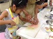 母親に教わりながら、慣れない手つきで「愛鯛」の切り身に包丁を入れる女の子＝２０１５年８月９日（日本財団撮影）