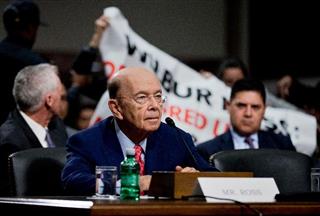 上院の公聴会で証言するウィルバー・ロス次期商務長官。後方ではロス氏に抗議する人たちが幕を広げた＝１８日、ワシントンの連邦議事堂（ＡＰ）