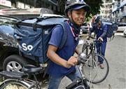宅配用の箱を背負い、市内各地に向けて自転車で出発する配達仲介サービスの配達員（共同）
